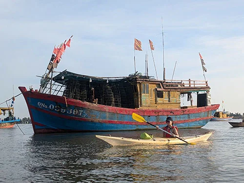 A tourist in a single kayak rowing by a big wooden fishing boat