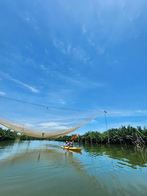 a double kayak with tourists rowing under an elevated fishnet under a blue open sky